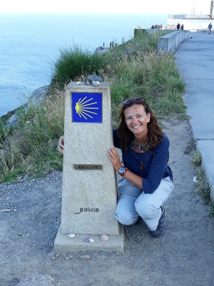 Barbara sorridente accanto al pilastro Km 0.000 di Galicia, con la conchiglia gialla del Cammino di Santiago, sullo sfondo il mare